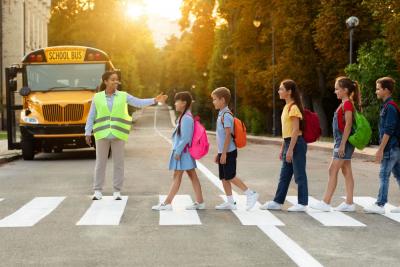 Traffic Safety: A woman in a crossing guard vest guides a group of children with colorful backpacks cross the road at a crosswalk.