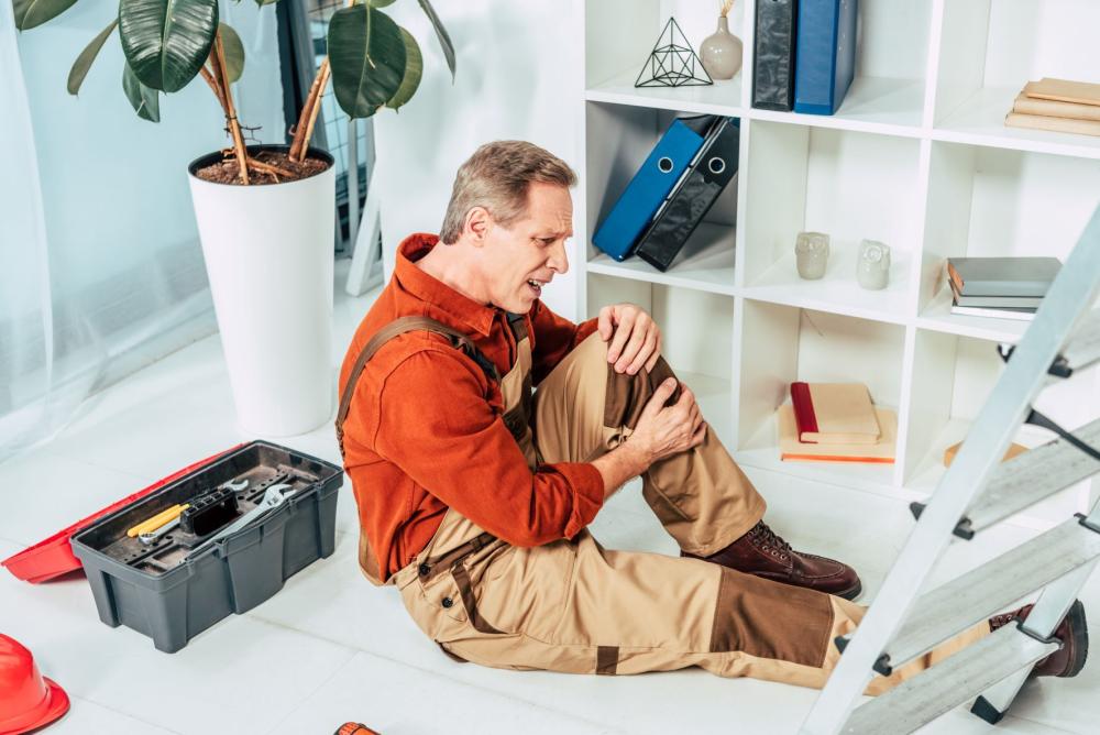 Repairman sitting on floor and holding injured knee surrounding by equipment in office